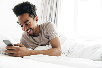 Wall Mural - Photo of smiling african american man using mobile phone in white bed