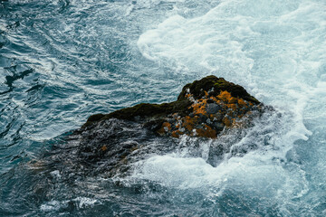 Mossy stone in azure water of mountain river close-up. Blue nature background with boulder with moss in turquoise water of mountain creek. Full frame of sea surf. Stone in ocean surf. Backdrop of tide