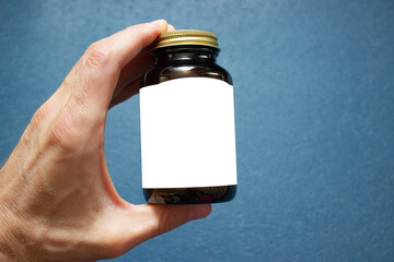 Wall Mural - A male hand holds a brown medical glass bottle with a white label for your text and a metal cap on a blue background.