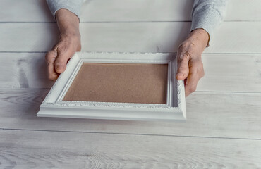 Wall Mural - Senior wrinkled hands holding white photo frame. Elderly woman looking at the picture, light wodden background. Empty template, mock-up. Happy memories, sadness, nostalgia concept.