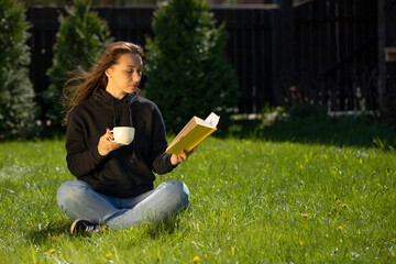 Attractive brunette model with long hair dressed in black hoodie sitting on grass reading book with cup of coffee on nice summer day. Relaxed vacation concept copy space. Distance Education Concept