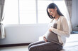 © Have a nice day  - beautiful asian pregnant woman smiling and hands rubbing firmly on her baby stomach, joyfully and happiness, sitting on the white bed relaxing and resting in the bedroom with light shining from window