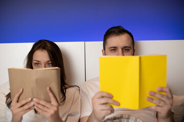 Beautiful young couple laying in bed with paper books in hands, girl and guy reading hiding behind open books, man and woman spend time together before bedtime, book store advertisement, bool lovers