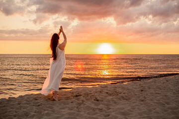 Happy young woman in a long white dress looking at the sunset on empty sand beach with her hands up. Freedoom, vacation, meditation