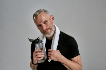 Middle aged muscular man in black t shirt looking at camera holding bottle of water and towel around his neck, posing in studio over grey background