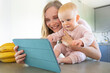 © Mangostar - Happy joyful mom and baby daughter talking to family in kitchen, using tablet for video call, smiling at screen together. Child care or online communication concept