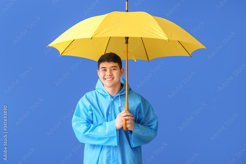 Young Asian man in raincoat and with umbrella on color background