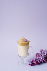 Beautiful dalgona drink a frothy coffee in a transparent mug and flowers lilac to purple Il purple background. Next to the mug is a marshmallow and a branch of lilac.