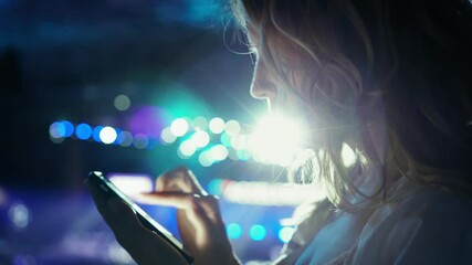 Attractive young woman with long hair writes message using a smartphone and smiling on the background of twinkling neon lights. Side view, close-up