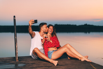 Wall Mural - Young couple sitting on the dock and taking selfies during summer evening