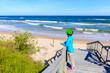© pkazmierczak - Young woman cyclist standing on steps to beautiful white sand beach with blue sea near Kolobrzeg, Baltic Sea coast, Poland