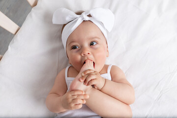 Wall Mural - baby girl 6 months old lies in a crib in the nursery with white clothes on her back and laughs, looks at the camera, baby's morning, baby products concept