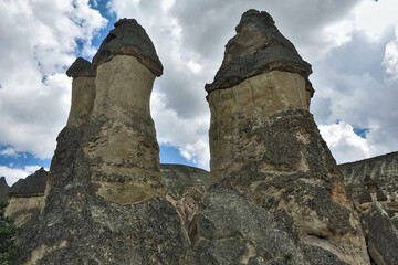 The amazing landscape of Cappadocia. Against the background of the sky with picturesque clouds towering cylindrical cliffs of tufa with dark conical basalt peaks. Rocks are like fantastic mushrooms. 