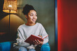 © BullRun - Smiling pretty african american girl sitting  with book at comfortable home interior, cheerful young dark skinned woman enjoying free time at apartment making notes of planning in textbook .