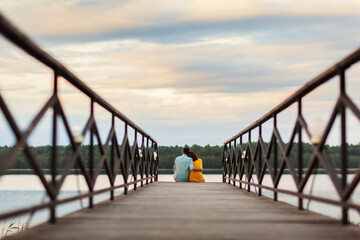 Back perspective view of young loving couple sitting on wooden pier, looking at beautiful river view and embracing warmly on summer day