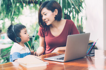 Wall Mural - Young asian mother and son using laptop computer for study and learning together at home, boy wearing headphone for e-learning with distancing, teacher or mom support child, education concept.