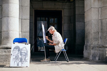 Old Male Street Painter Drawing Portrait Of Tourist