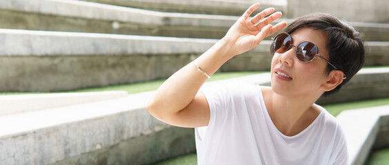 Stylish, middle aged Asian woman wearing 100% UV light protection sunglasses, raise her hand to block out bright glare and sunlight from outside to avoid ultraviolet rays overexposure. 