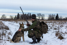 Male Hunter In Camouflage Clothes Walking On The Snow Field With Hunting Rifle During A Hunt, Dog Follows Him, Foggy Weather