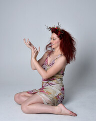 Poster - full length pose of red hair girl wearing floral fairy dress and headdress. sitting pose on grey studio background.