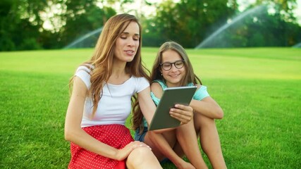 Wall Mural - Mother and daughter laughing in park. Woman and girl talking at camera on tablet