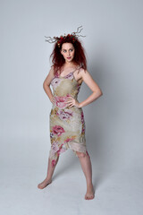 full length portrait of red haired girl wearing floral dress and headdress. standing pose on grey studio background.