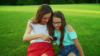 Wall Mural - Woman and girl sitting on grass. Mother and daughter looking video on smartphone