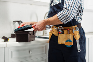 Cropped view of plumber in overalls opening toolbox on worktop in kitchen