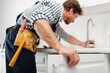 © LIGHTFIELD STUDIOS - Selective focus of plumber in tool belt repairing kitchen faucet