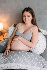 Wall Mural - pregnant girl model woman sitting on bed in tracksuit near pink peonies with hug pillow