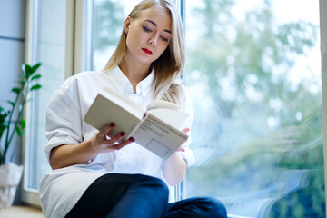 Confident blonde female marketing manager resting on break sitting on window sill with favorite book, concentrated pretty student fond of reading analyzing information preparing for examinations