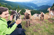 © anatoliy_gleb - Close up of laughing brunette woman sitting on grass near group of mammal animals, mountain scenery with forest on background. Rocky mountain goats in warm sunny summer day, cloudy sky.