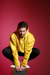 Wall Mural - A young man of 25-30 years old in a yellow sweatshirt emotionally standing on an old music column on pink wall background. 