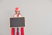 Cute Little Girl In Superhero Costume Holding Empty Chalkboard With Space For Your Design Against Grey Background