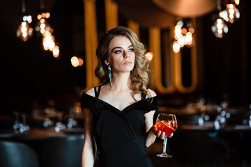 Beautiful blonde, young girl holding an aperol spritz. Cocktail aperol spritz in a glass. Portrait of a young girl in a restaurant. Portrait of a girl. Young girl in a restaurant with a glass.