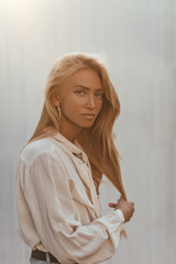 Tanned blonde blue-eyed long-haired woman in light beige cotton blouse looks into camera and poses outside near white wall.