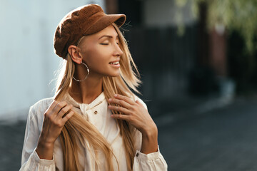 Wall Mural - Happy calm young tanned blonde woman in brown cap and white shirt smiles sincerely and poses outside.