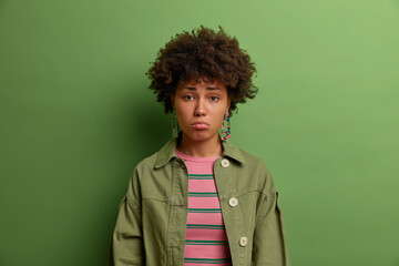 Wall Mural - Sad depressed ethnic woman looks with resentful expression, looks dissatisfied and gloomy, worries about difficulties, wears green jacket, poses indoor. People and negative emotions concept.