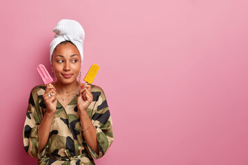 Sticker - Portrait of thoughtful African American woman holds strawberry and mango ice cream, wears domestic clothes, thinks what better to eat, looks aside, isolated on pink background, blank copy space aside