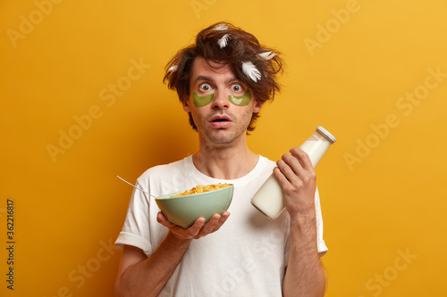 Photo of young man eats cornflakes with milk for breakfast, stares with bugged eyes as hears shocking news in morning, has healthy nutrition, cares about eye skin, wears patches for reducing wrinkles