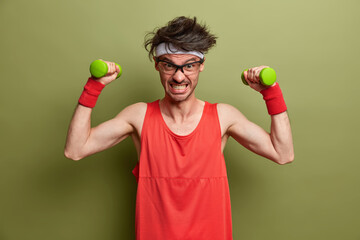 Funny sportsman puts all efforts in lifting heavy dumbbells, clenches teeth with force, does home exercises, enjoys bodybuilding, stands motivated against green background. Healthy lifestyle concept