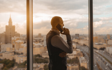 Wall Mural - Silhouette of a hispanic businessman talking on the phone while standing near a panoramic window of a luxury office skyscraper with a roadway and a high-rise with a spire outside, beautiful sunset