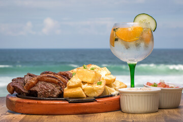 Picanha, a traditional cut of Brazilian meat, served with fried cassava on a hot plate. In the background there's a tropical drink and the beach