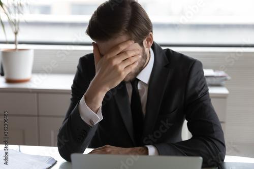 Frustrated male employee sit at desk work on laptop feel stressed with company bankruptcy news, disappointed businessman distressed disappointed with corporate business failure or money loss