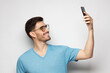 © Damir Khabirov - Young handsome man in blue tshirt holding phone and taking selfie photo, isolated on gray background
