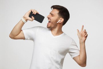 Young man in white t-shirt listening to music in wireless earphpones and singing in online karaoke app using smart phone, isolated on gray background