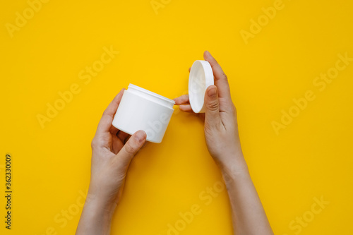 Human hands opening a white blank body cream jar on clean yellow background.