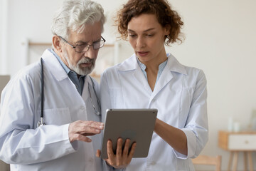 Concentrated man and woman doctors in white medical uniforms look at tablet screen discuss patient anamnesis or checkup results together, diverse GP talk brainstorm make decision using pad gadget