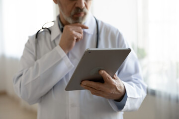 Wall Mural - Crop close up of mature male doctor physician in medical uniform look at tablet screen make decision, pensive man GP or therapist work use modern pan gadget in hospital, medicine technology concept