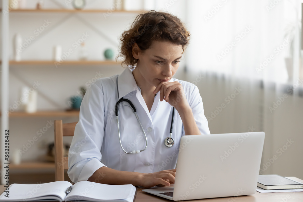 Concerned young female Caucasian doctor in white medical uniform sit at desk in hospital work on laptop, focused woman nurse or GP busy fill anamnesis on computer gadget, medicine, technology concept - obrazy, fototapety, plakaty 